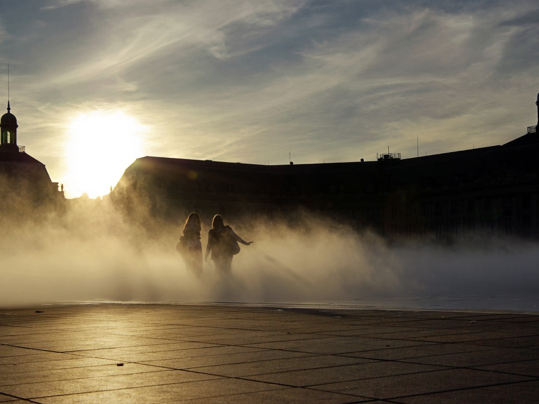 Two figures walking through the mist over the water mirror at Bordeaux, the classical frontage of the place de la Bourse dark against the low sun.