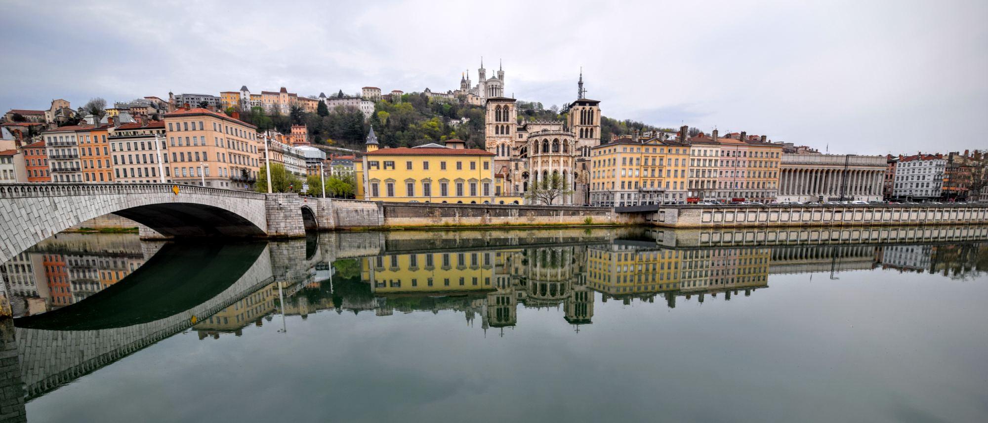 The Lyon waterfront across flat water: a long terrace of pale quayside buildings with the wooded hill of Fourvière rising behind them.