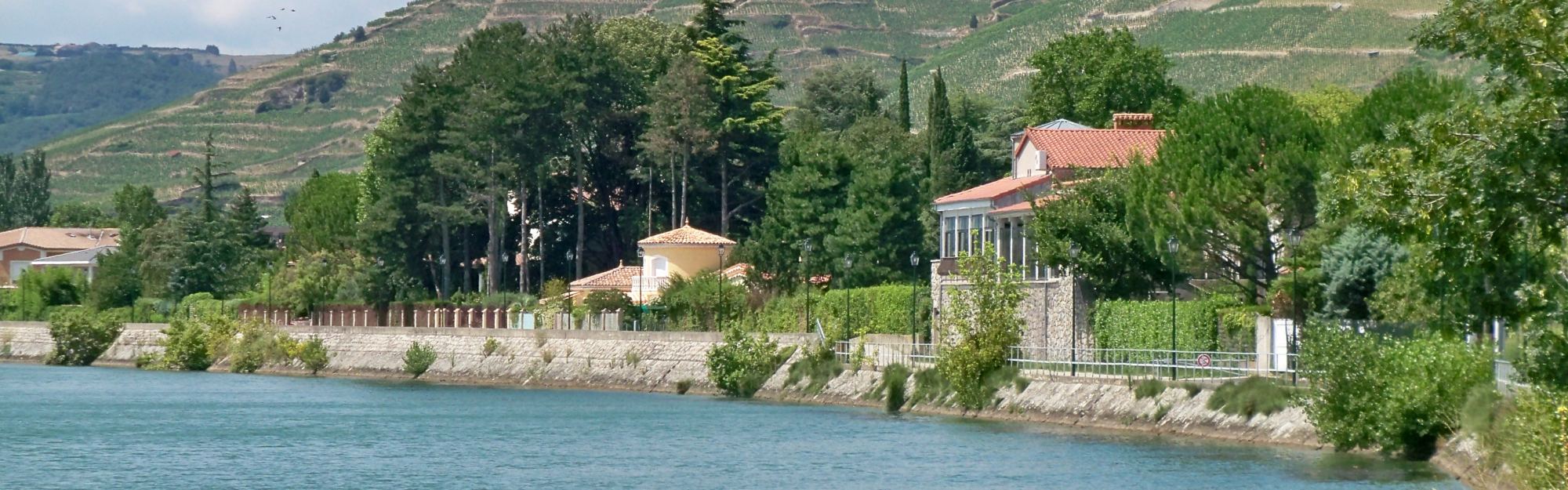 The Rhône running past a stone quay wall at Tain-l’Hermitage, with the terraced vineyard hill rising directly behind the riverside houses.