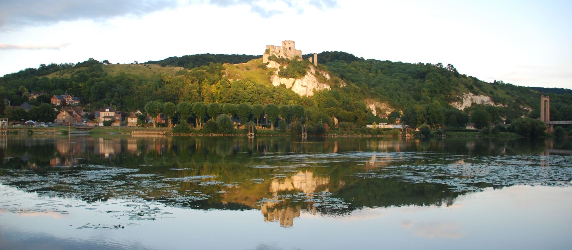 Château Gaillard standing on the chalk ridge above the Seine at Les Andelys, the ruin and the wooded slope reflected in still water at sunset.